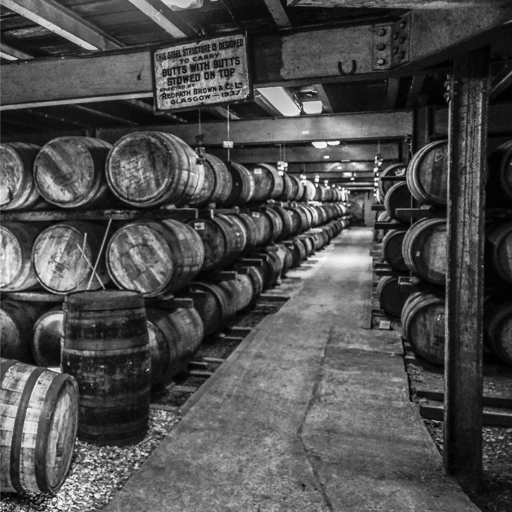 Black and white image of a warehouse. bourbon barrels. old sign that reads "This steel structure is designed to carry butts with butts sttowed on top, erected by redpath brown & co glasgow 1937"